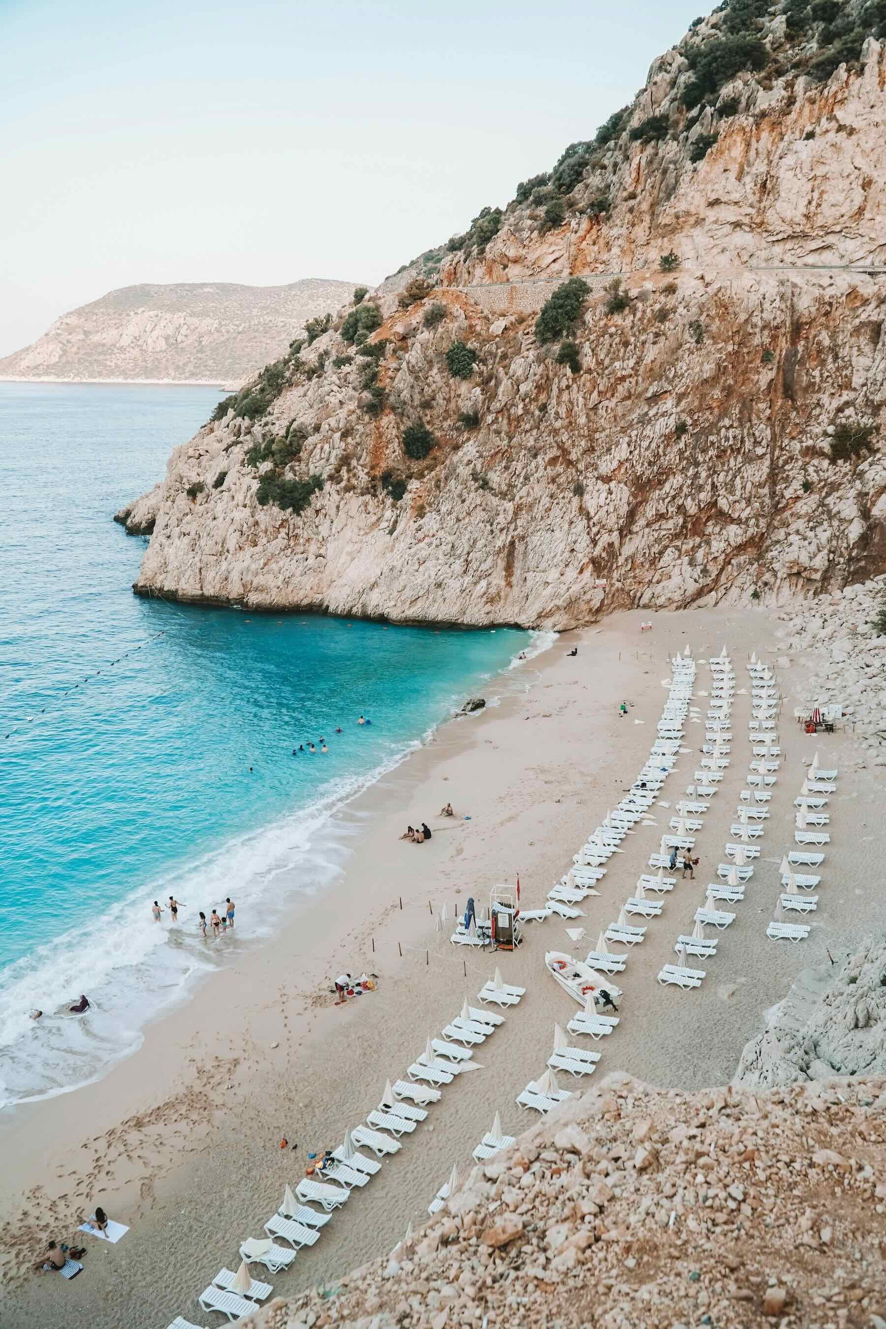 View of a beach in Antalya, Turkey. There are people on the beach and a line of white sun loungers facing the sea. There is a cliff in the background.