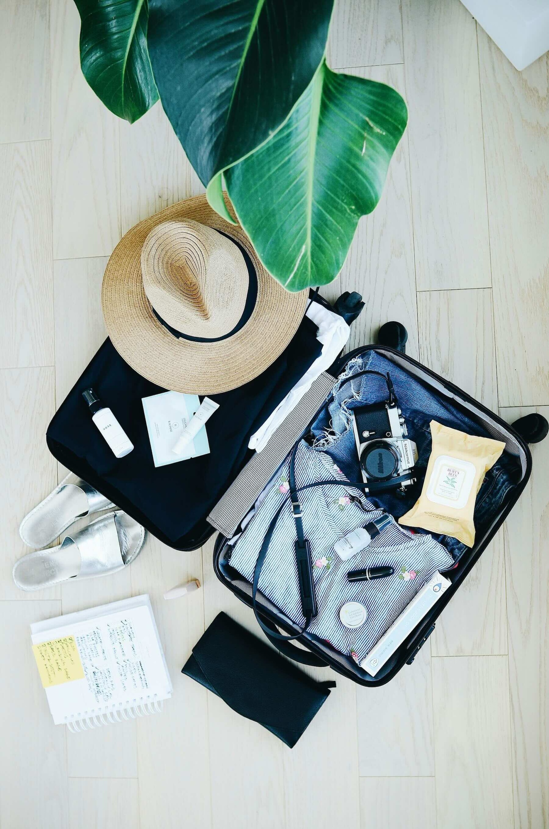 An open suitcase lies on the floor with travel items in it including a wide brim sun hat, a camera, a pair of sandals a notebook and folded clothes.