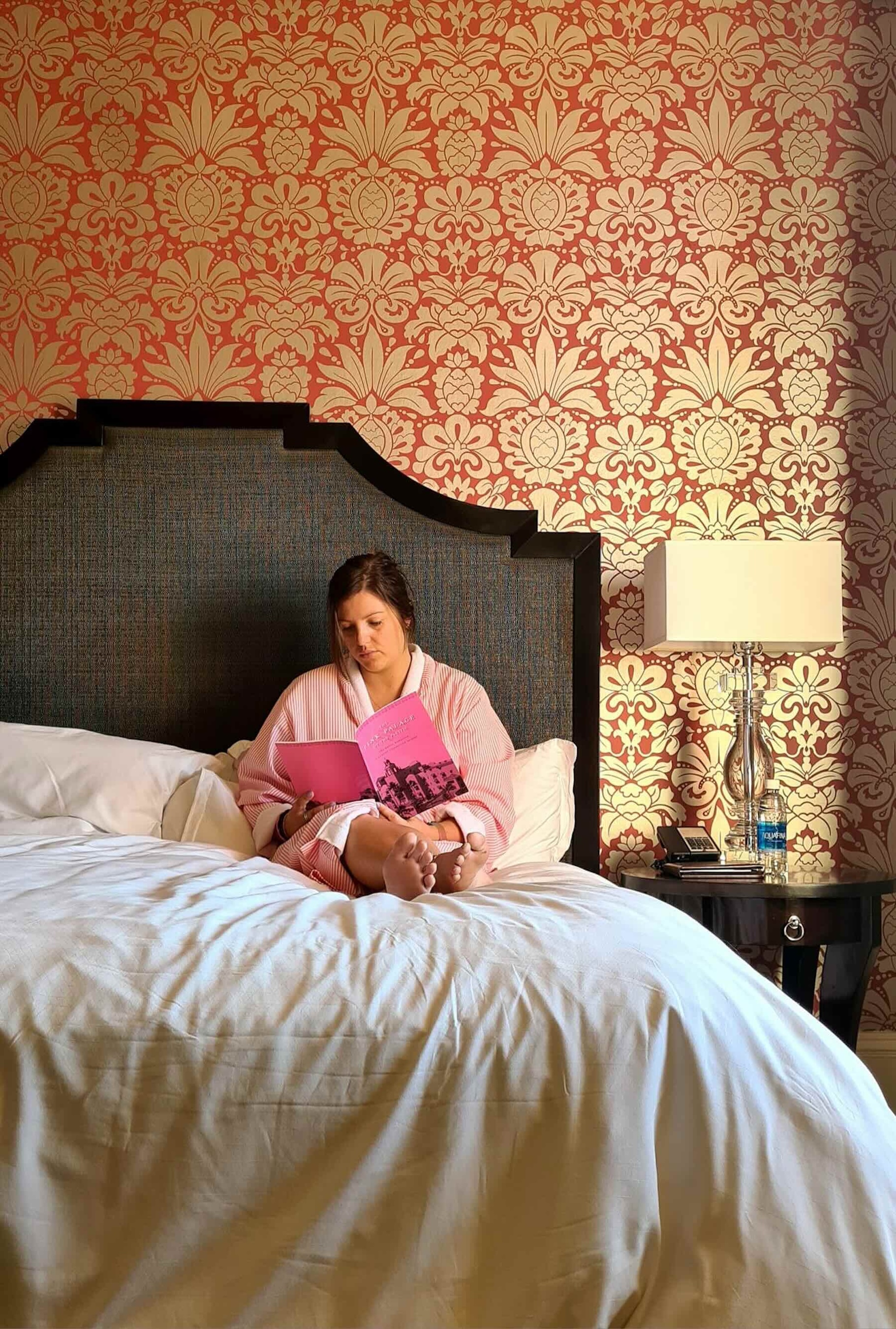 Jade, the owen of Roast and Roam, sits on a large hotel bed reading a book. There is a statement wall with red and gold wallpaper behind her. Next to the bed is a bedside table with a lamp and telephone.