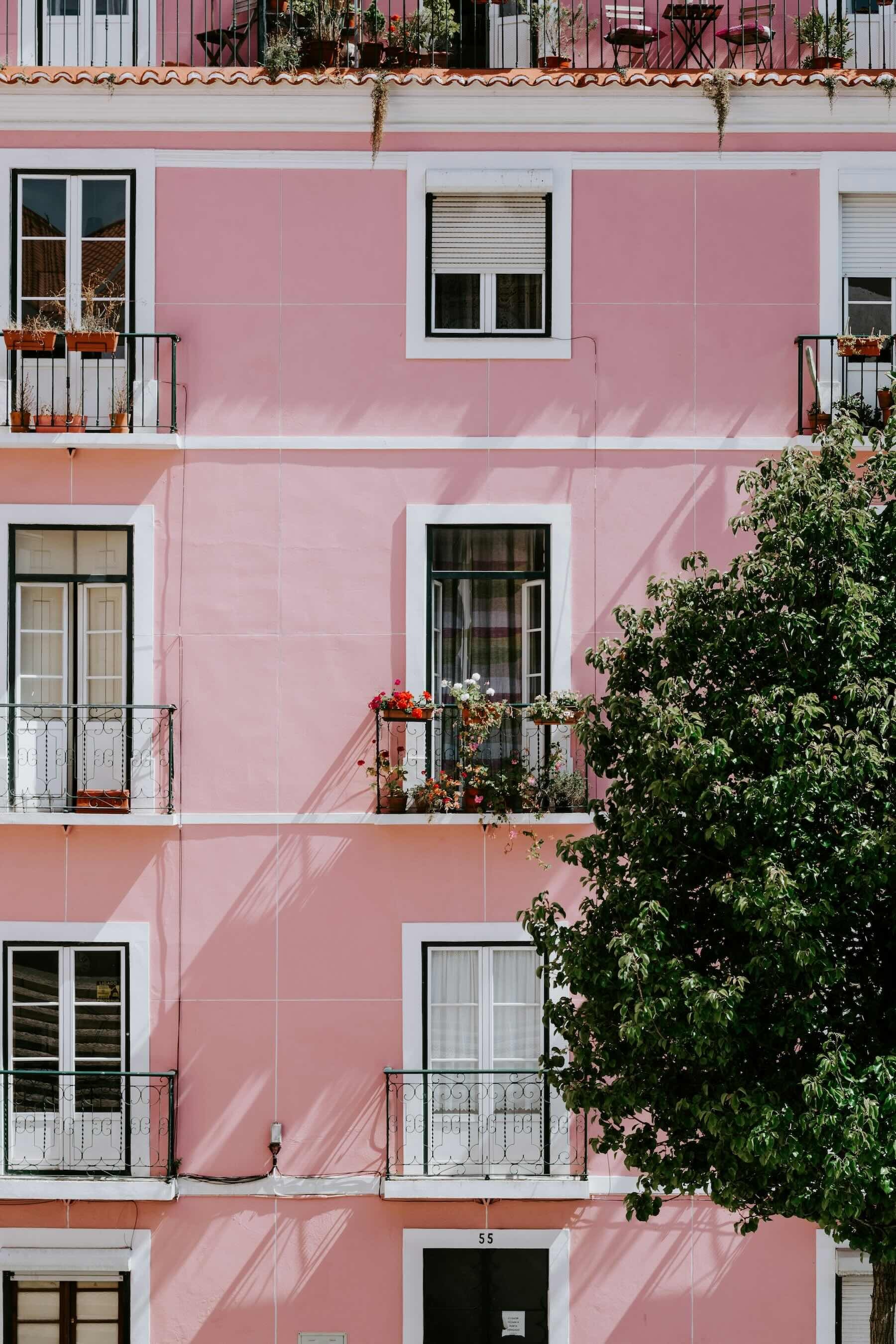 A four storey block of apartments in Lisbon, Portugal, the wall is pink, the window frames and door frames are white, some of the doors have balconies with flower boxes hanging from them.