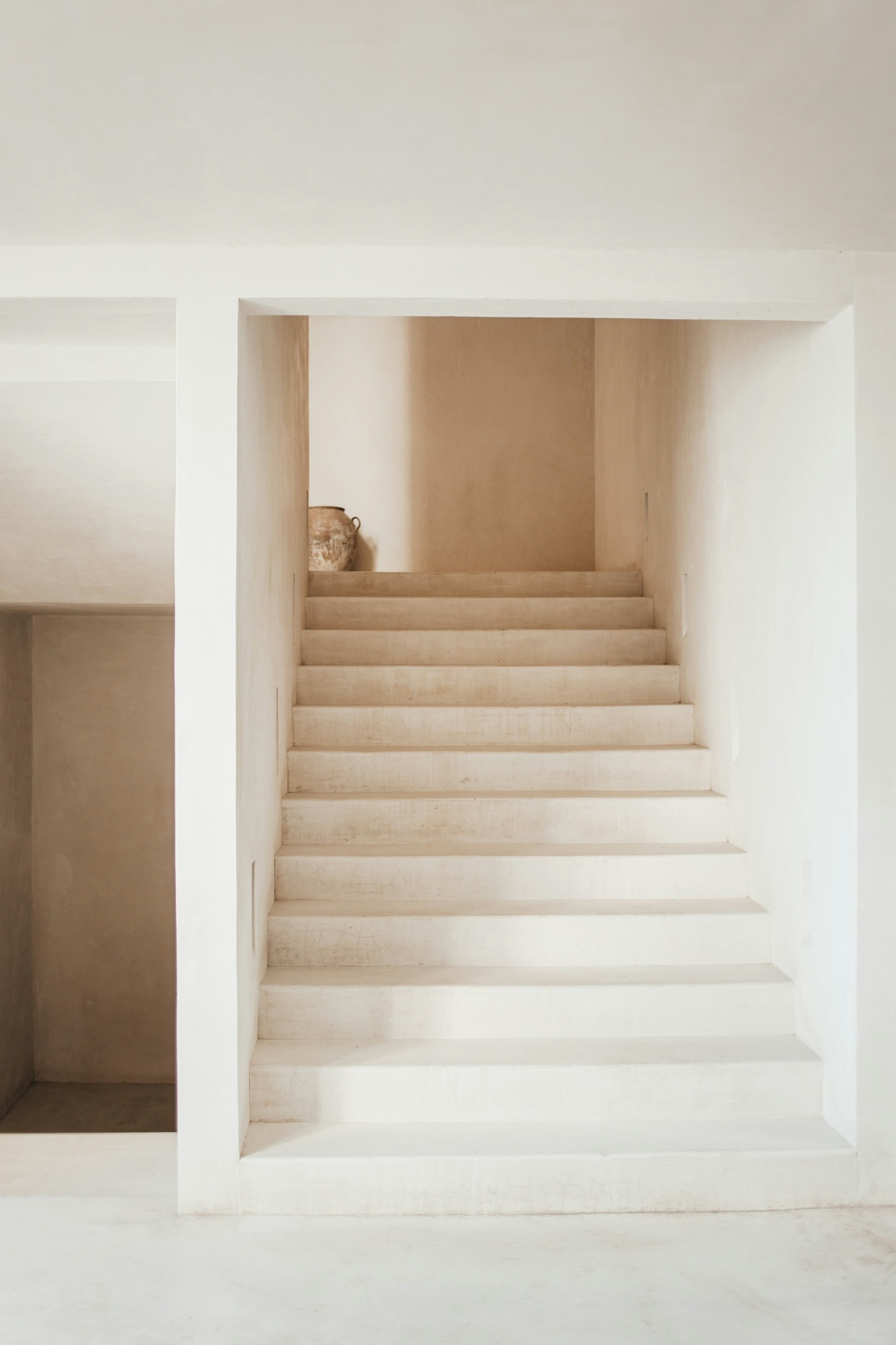 An internal stairwell. The walls and stairs are all off-white concrete. At the top of the stairs is a large, rustic, terracotta vase.